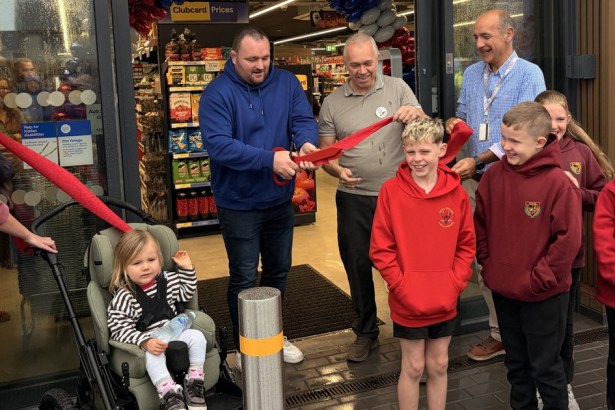 Former Wales rugby captain Ken Owens, in a blue hoodie, cuts a red ribbon with giant scissors at the entrance to Tesco Express in Tumble, beside store manager Martin Lewis, with children and staff looking on