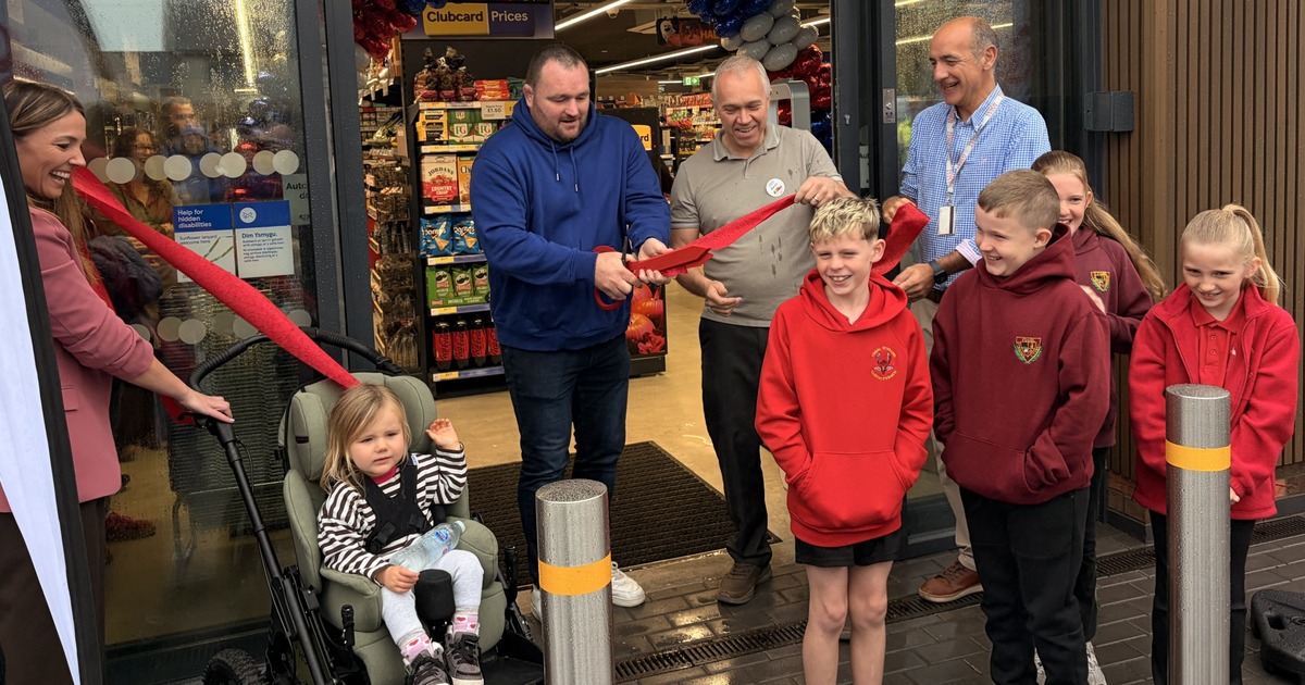 Former Wales rugby captain Ken Owens, in a blue hoodie, cuts a red ribbon with giant scissors at the entrance to Tesco Express in Tumble, beside store manager Martin Lewis, with children and staff looking on