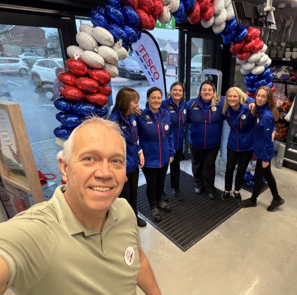 Store manager Martin Lewis takes a selfie in front of six smiling staff in blue Tesco jackets standing under a red, white and blue balloon arch at the store entrance
