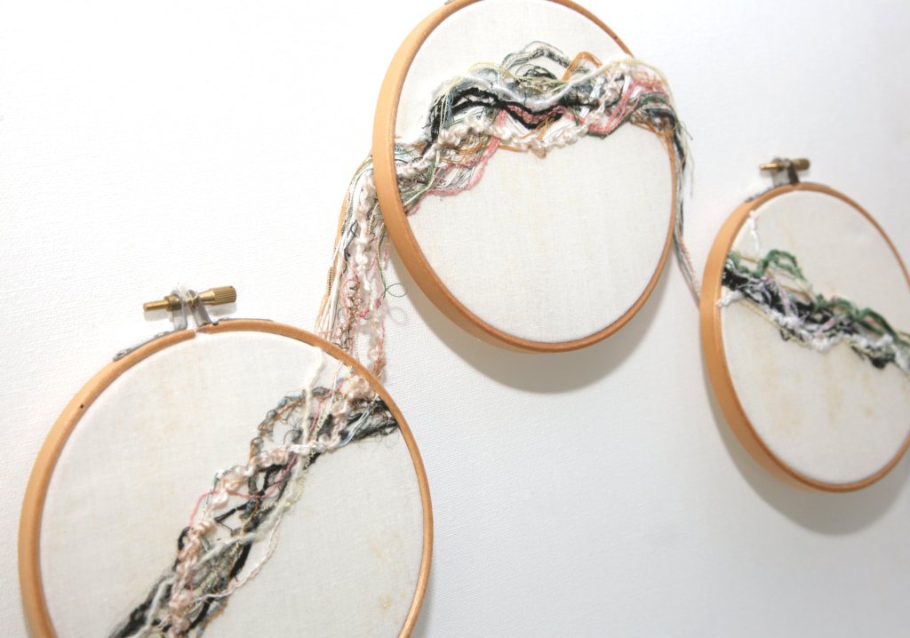 Three wooden embroidery hoops on a white wall, joined by a trail of tangled pink, black, green and cream threads