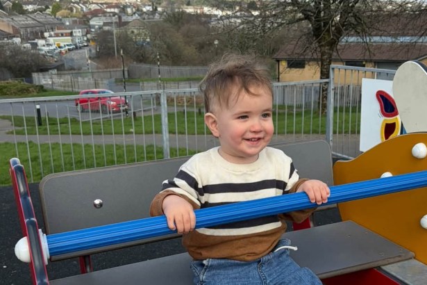 A smiling toddler in a striped jumper holding a blue bar on playground equipment