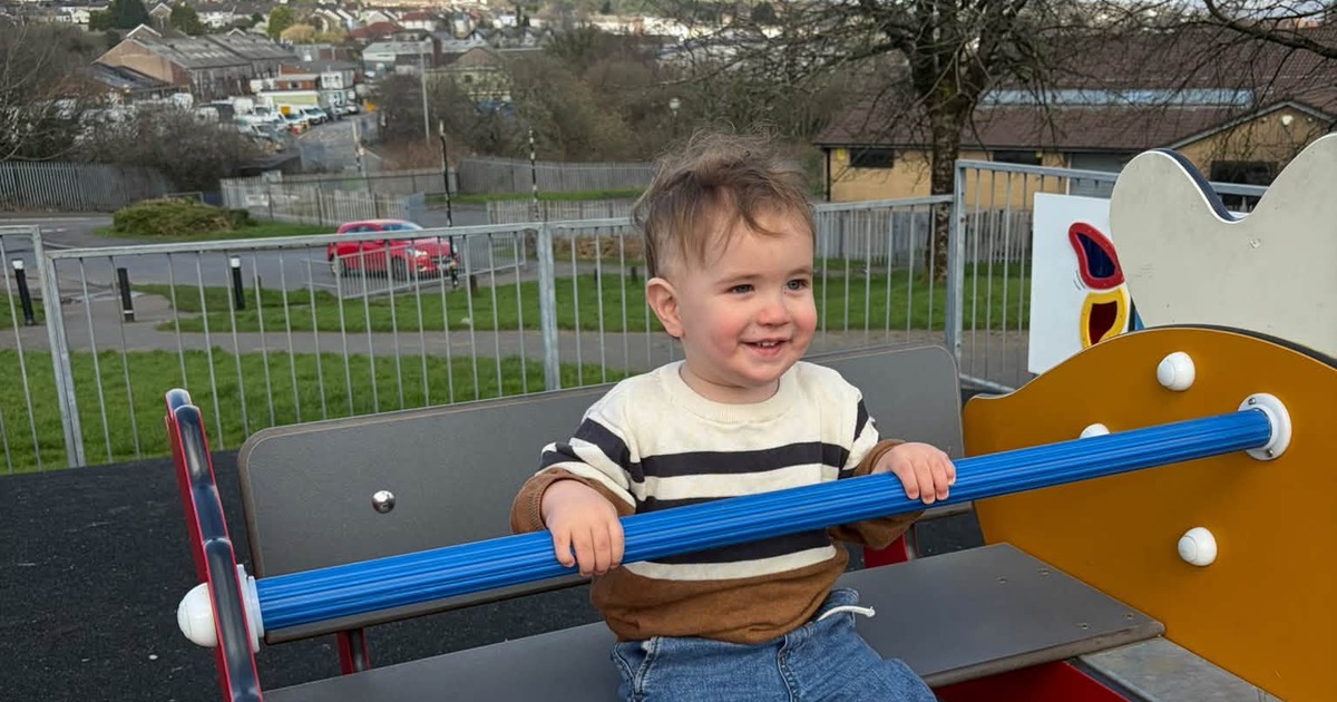 A smiling toddler in a striped jumper holding a blue bar on playground equipment