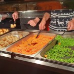 Volunteers in Christmas jumpers serving roast potatoes, carrots, peas and sprouts from a servery at Together at Christmas in the Brangwyn Hall