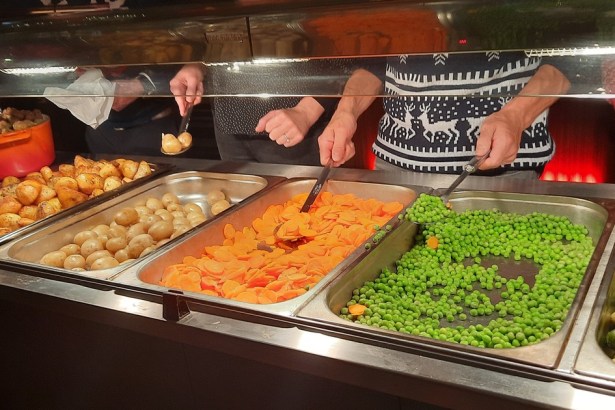 Volunteers in Christmas jumpers serving roast potatoes, carrots, peas and sprouts from a servery at Together at Christmas in the Brangwyn Hall