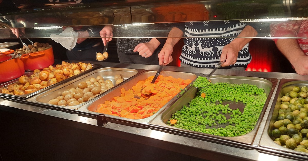 Volunteers in Christmas jumpers serving roast potatoes, carrots, peas and sprouts from a servery at Together at Christmas in the Brangwyn Hall