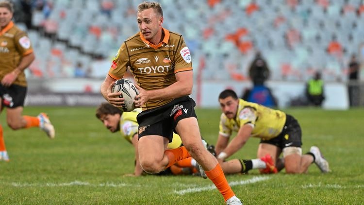 A Toyota Cheetahs player in an orange and black kit runs with the ball as two defenders in yellow are left on the ground behind him