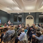 Residents seated in rows in a high-ceilinged room listen to a speaker with a microphone beside a panel table, with a screen listing the evening's speakers