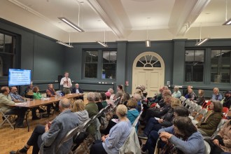 Residents seated in rows in a high-ceilinged room listen to a speaker with a microphone beside a panel table, with a screen listing the evening's speakers