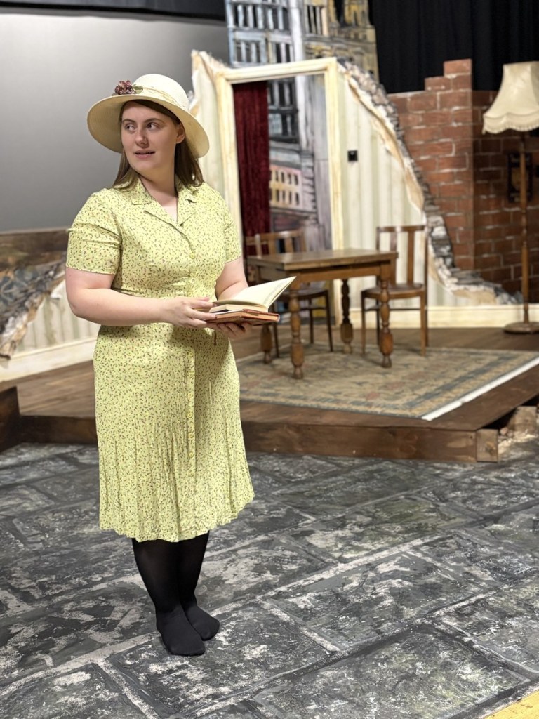 An actress in a yellow dress and straw hat reads from a book on the Llareggub set in Swansea Little Theatre's Under Milk Wood