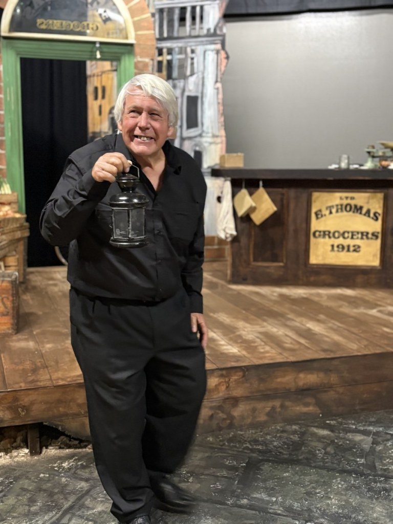 A white-haired actor dressed in black holds up a lantern and grins on the Llareggub set in Swansea Little Theatre's Under Milk Wood