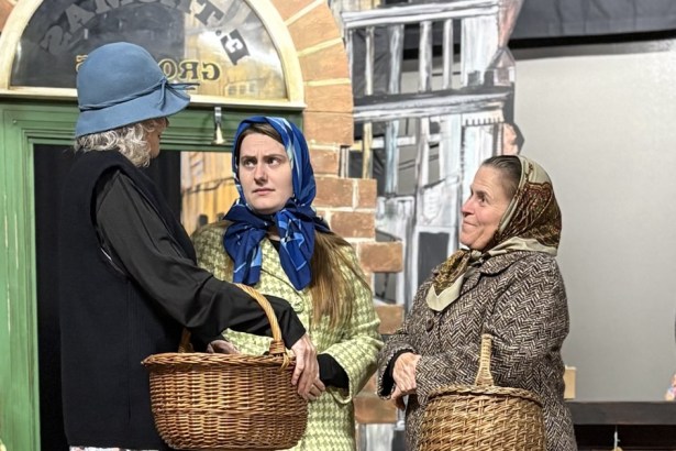 Three women in headscarves and hats carrying wicker baskets outside the E Thomas Grocers shopfront in Swansea Little Theatre's Under Milk Wood