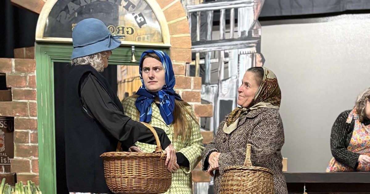 Three women in headscarves and hats carrying wicker baskets outside the E Thomas Grocers shopfront in Swansea Little Theatre's Under Milk Wood