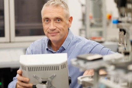 A grey-haired man in a blue checked shirt holding a robot control panel among automation equipment in a lab