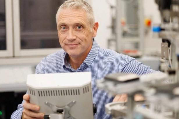A grey-haired man in a blue checked shirt holding a robot control panel among automation equipment in a lab