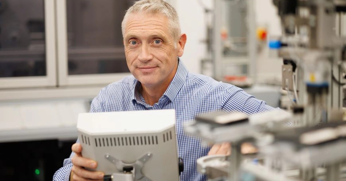 A grey-haired man in a blue checked shirt holding a robot control panel among automation equipment in a lab