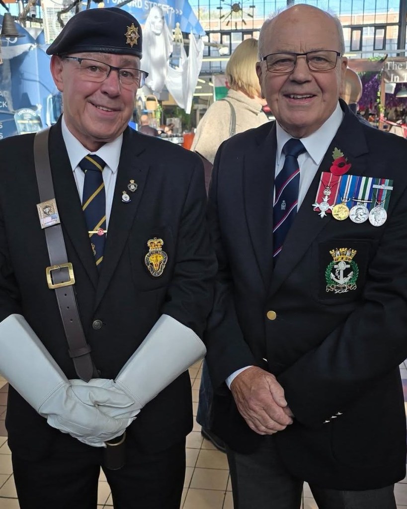 Two veterans in navy blazers, one in a beret and white gauntlets, the other wearing medals and a poppy, smiling in Swansea Market.