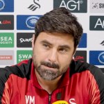 Swansea City head coach Vitor Matos, in a red and black training top, at his press conference in front of a sponsor board