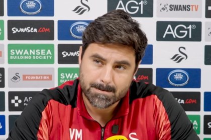 Swansea City head coach Vitor Matos, in a red and black training top, at his press conference in front of a sponsor board