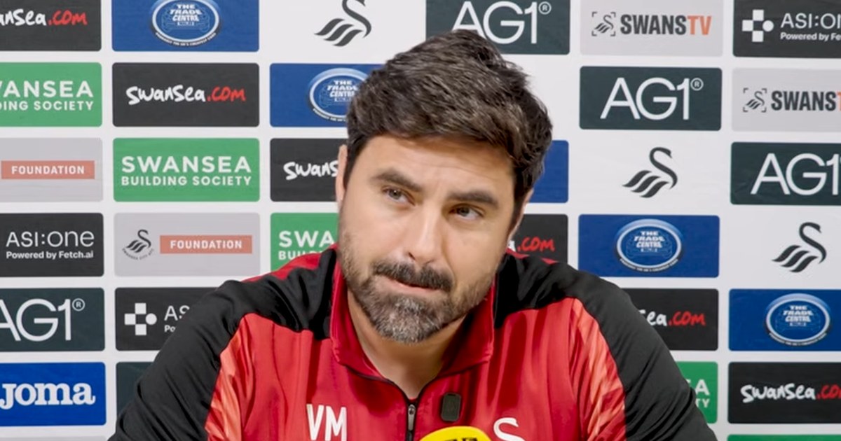 Swansea City head coach Vitor Matos, in a red and black training top, at his press conference in front of a sponsor board