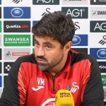 Swansea City head coach Vitor Matos, in a red and black training top, speaking at his press conference in front of a sponsor board