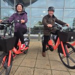 Two councillors in cycle helmets sitting on orange Voi e-bikes with front baskets outside a glass-fronted building, a National Express coach behind