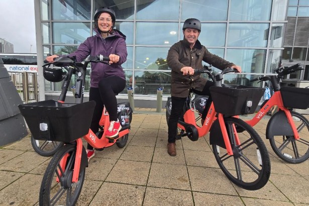 Two councillors in cycle helmets sitting on orange Voi e-bikes with front baskets outside a glass-fronted building, a National Express coach behind
