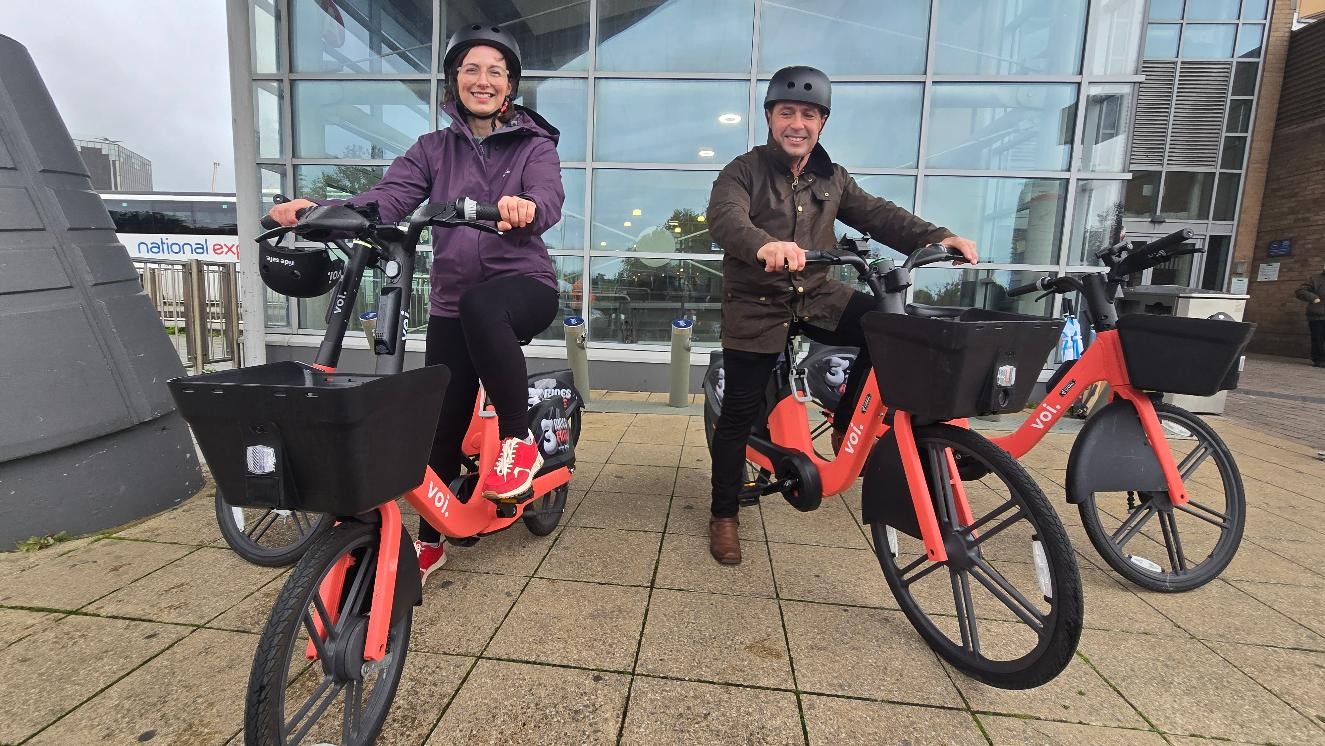 Two councillors in cycle helmets sitting on orange Voi e-bikes with front baskets outside a glass-fronted building, a National Express coach behind