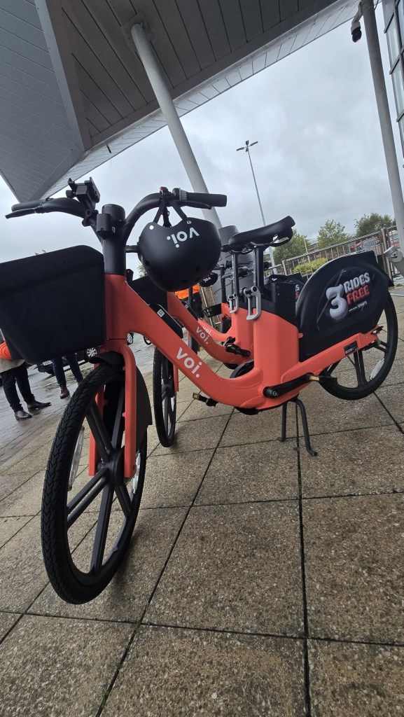 Two orange Voi e-bikes with front baskets and a black Voi helmet hanging from the handlebars, parked under a canopy