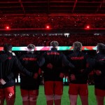 Wales players in black Cymru tops with arms linked, seen from behind, facing a packed Principality Stadium lit in red