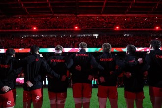 Wales players in black Cymru tops with arms linked, seen from behind, facing a packed Principality Stadium lit in red
