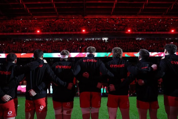 Wales players in black Cymru tops with arms linked, seen from behind, facing a packed Principality Stadium lit in red