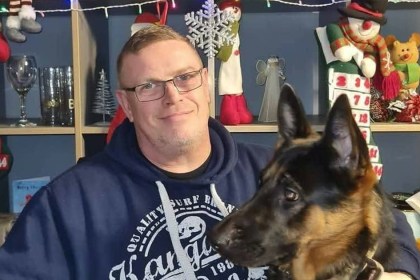 Wayne Bowron, a man in glasses and a navy hoodie, smiling beside a German shepherd dog in front of Christmas decorations