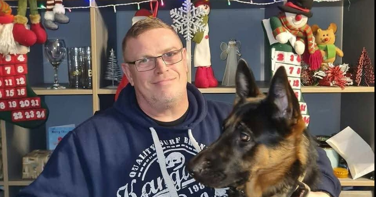 Wayne Bowron, a man in glasses and a navy hoodie, smiling beside a German shepherd dog in front of Christmas decorations