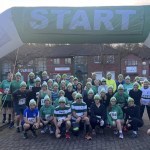Dozens of runners in green-and-white bobble hats and race numbers posing under an inflatable green START arch