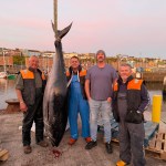Four fishermen in waterproofs and a man in a grey T-shirt stand beside a bluefin tuna hung by its tail from a hoist on the quay at Milford Haven at dusk