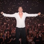 Will Parfitt in a white shirt with arms outstretched on stage, in front of a crowd holding up phone lights