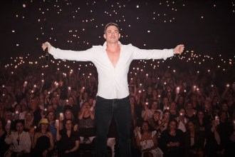 Will Parfitt in a white shirt with arms outstretched on stage, in front of a crowd holding up phone lights