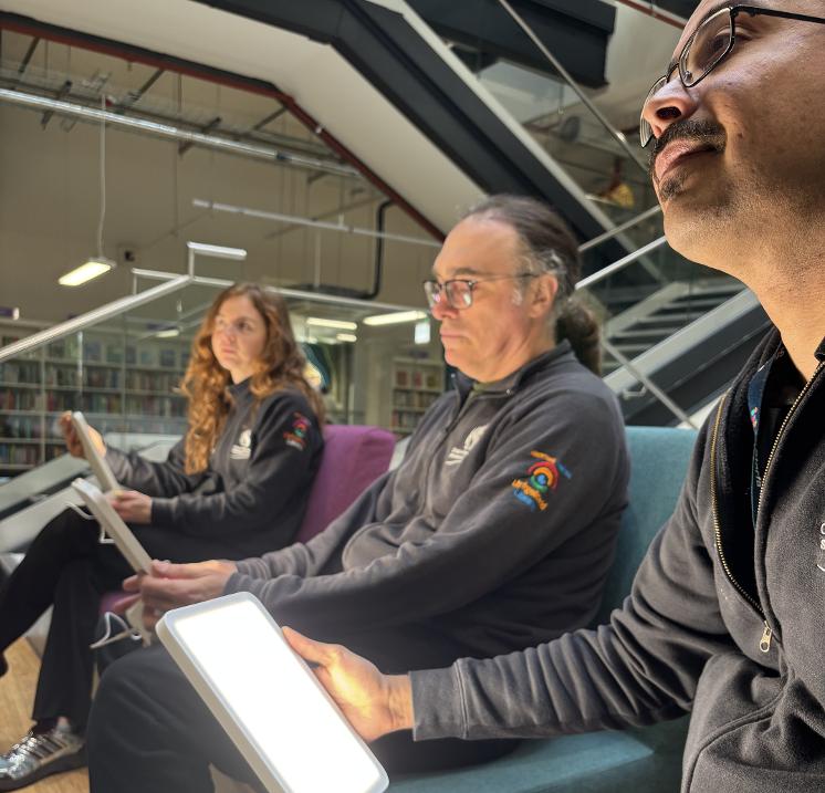 Three library staff members sitting on sofas with lit daylight lamps on their laps