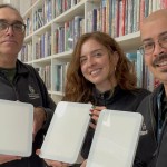 Three Swansea library staff members holding up white daylight lamps in front of bookshelves