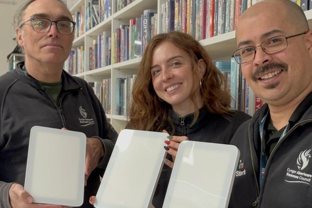 Three Swansea library staff members holding up white daylight lamps in front of bookshelves