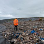 An NRW officer in orange walks across exposed waste at Withyhedge Landfill with gulls overhead and capped slopes behind