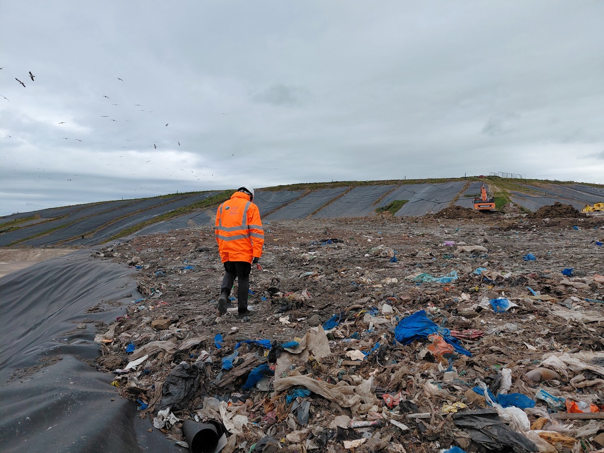 An NRW officer in orange walks across exposed waste at Withyhedge Landfill with gulls overhead and capped slopes behind