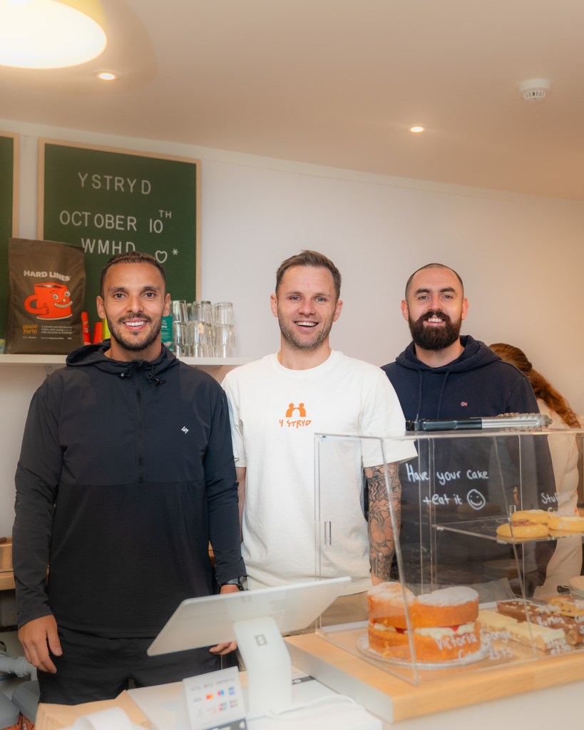 Three men smile behind the cake counter at Y Stryd, with a World Mental Health Day sign behind them