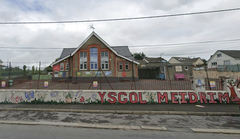 Ysgol Meidrim's red-brick gabled school building behind red railings, with the school's name painted in large red letters along a front wall decorated with flowers, butterflies and toadstools