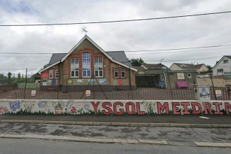 Ysgol Meidrim's red-brick gabled school building behind red railings, with the school's name painted in large red letters along a front wall decorated with flowers, butterflies and toadstools