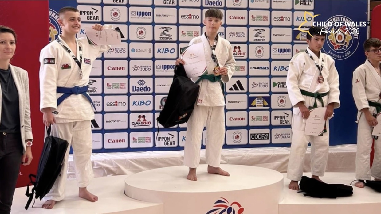Young judo players in white judogi on a medal podium in front of a sponsor board, holding certificates