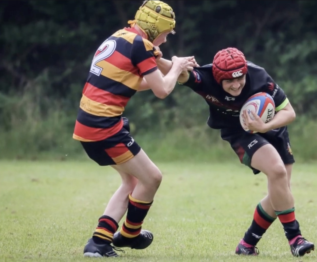A young rugby player in a red scrum cap carrying the ball into a tackle from an opponent in a striped shirt