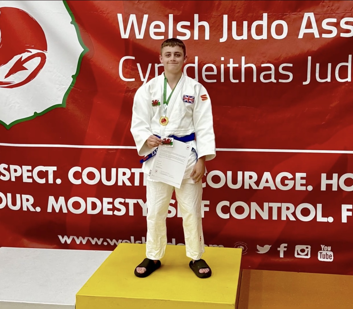 Zac Thomas in a white judogi with a gold medal and certificate, standing on the top step of a podium in front of a Welsh Judo Association banner