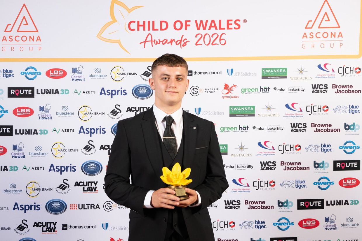 Zac Thomas in a black three-piece suit holding a yellow daffodil trophy in front of the Child of Wales Awards 2026 sponsor board